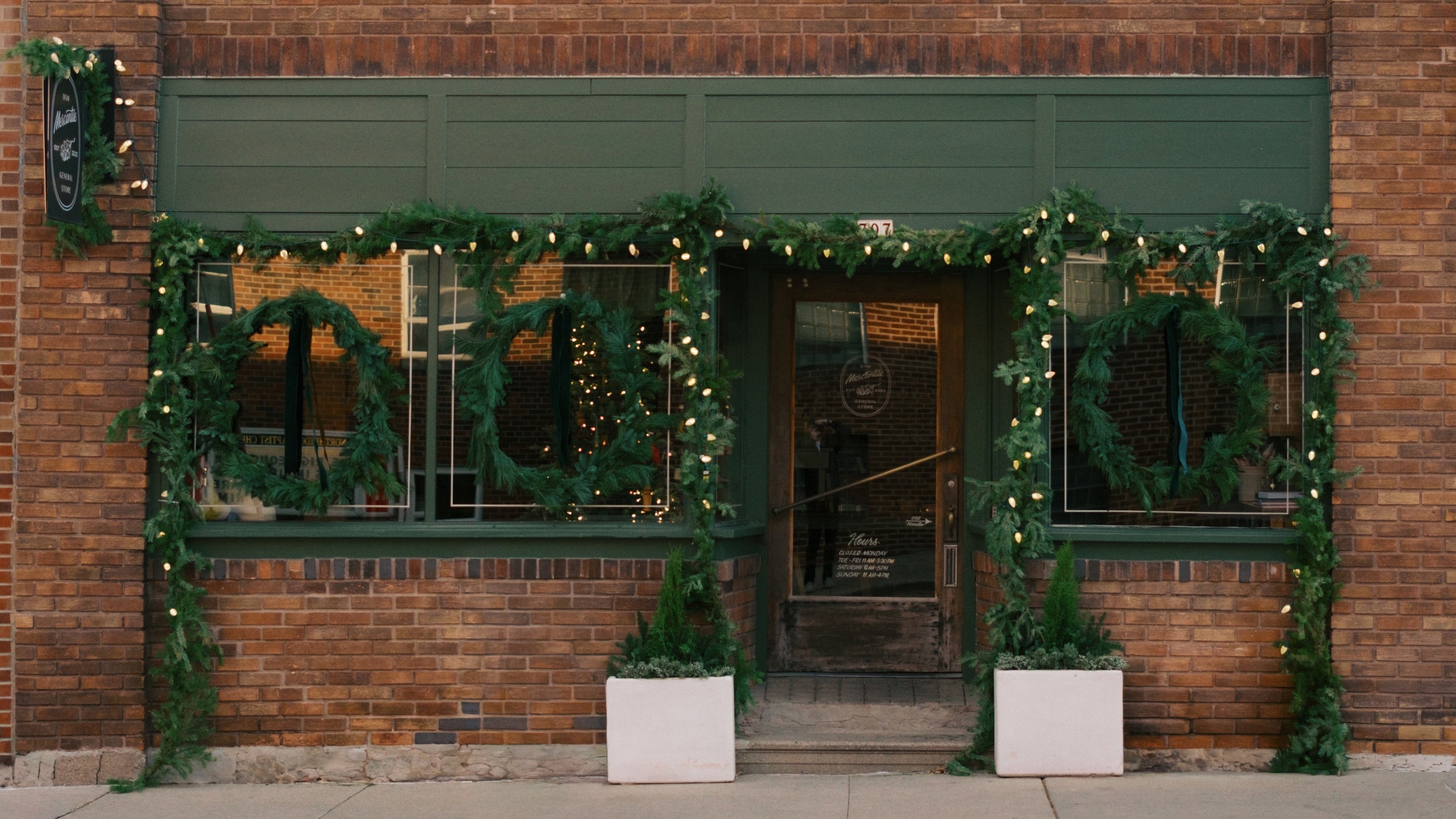 Brick building with 'Des Moines Mercantile' sign, decorated with greenery and lights.